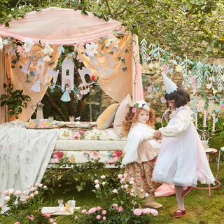 Two children in costume play by a garden party setup with flowers, a floral canopy-topped bench, enchanted fairy tale decorations, and the Princess Party Garland from Meri Meri.