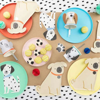 A playful assortment of dog-themed party decorations, including Meri Meri's Puppy Face with Droopy Ears Cups made from sustainable FSC paper adorned with cute puppy illustrations, soccer ball accents, and colorful macarons, arranged on a polka