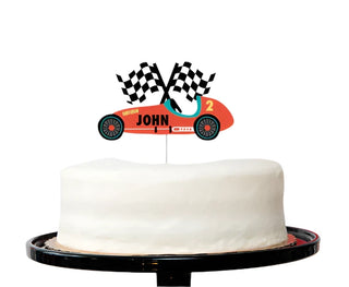 A white frosted cake on a black stand is decorated with the Race Car Custom Cake Topper from Merrilulu, featuring two checkered flags and the name "John" along with the number "2." This setup is perfect for a car-themed birthday party, complete with a red balloon in the background.