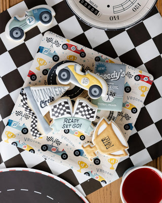 Decorated cookies themed around car racing, perfect for a birthday party, include a race car, checkered flags, a pit stop sign, and a car-themed book. They are set on a Race Pattern Plate by My Mind’s Eye atop a racing mat with black-and-white checkered and road track patterns—ideal for racing fans.