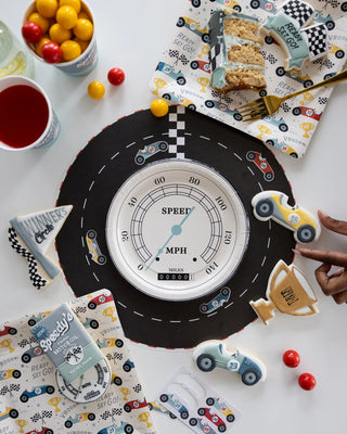 Overhead view of a car-themed birthday party setup with a Race Track Placemat by My Mind’s Eye, car-shaped cookies, checker-pattern napkins, yellow cup with red drink, and assorted candies.