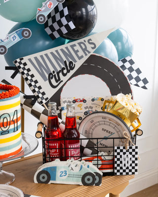 A racing-themed birthday party setup with a cake, balloons, two bottles of soda, popcorn, and various racing decorations. The table features a Race Track Placemat from My Mind’s Eye and a "Winner's Circle" sign, along with a toy race car sporting the number 23.