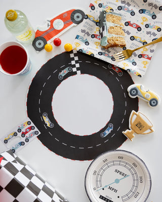 A race car-themed birthday party table setup with a circular Race Track Placemat from My Mind’s Eye, toy cars, themed napkins, plates, and cups. Cake slices sit on plates alongside beverages for an exciting celebration.