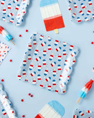 Flat-lay of playful 4th of July-themed stationery with My Mind's Eye Red White Blue Ice Pop Shaped Paper Guest Napkin designs, surrounded by scattered beads, evoking a cool, refreshing vibe against a pastel blue backdrop.