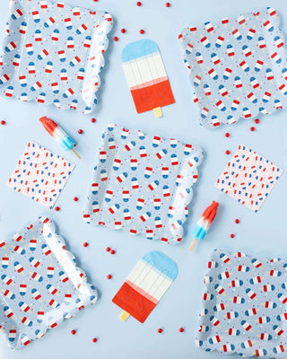 A playful flat lay with summer-themed items: fabric clothes with popsicle patterns, scattered red beads, and realistic toy popsicle figures alongside Red White Blue Ice Pop Shaped Paper Guest Napkins from My Mind's Eye on a pastel blue background, creating.