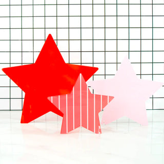 Three different-sized Red and Pink Acrylic Stars Decor - 4th of July by Kailo Chic in star-shaped cutouts with striped patterns are displayed against a checkered white and black background on a white surface, perfect for adding a touch of patriotic home decor to your 4th of July celebration.