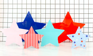Six colorful star-shaped objects, featuring solid colors, stripes, and a star pattern, are arranged in front of a grid-patterned backdrop on a white surface—a perfect touch for patriotic home decor. This is the Red and Pink Acrylic Stars Decor by Kailo Chic.