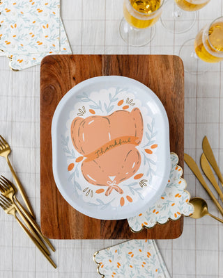 A table setting with a wood cutting board, My Mind’s Eye "Roasted Turkey" paper plates featuring a "Thankful" and pumpkin harvest design, gold cutlery, napkins with a matching pattern, and three glasses of yellow beverage.