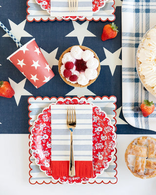 Overhead view of a festive table setting for a patriotic celebration, featuring star-patterned napkins, My Mind's Eye Stripped Scallop Plates, cutlery, a tart with whipped cream and raspberries, and scattered strawberries.