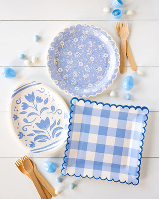 Two decorative Sweet Floral Lavender paper plates with a floral design, a blue checked napkin, and wooden cutlery arranged neatly on a white wooden surface with scattered blue stones around.