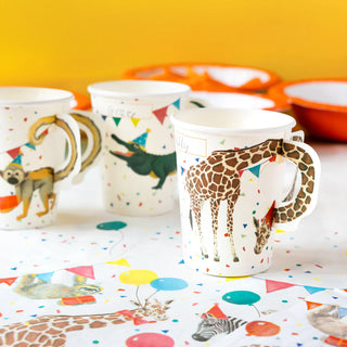 Three Safari Animal Paper Cups featuring illustrations of a giraffe and monkey, set on a table with a colorful, festive tablecloth and party decorations. These recyclable mugs by Talking Tables add an eco-friendly touch to your celebrations.