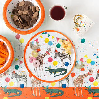 A party table with animal-themed decorations, including plates, bowls with snacks, Talking Tables' Safari Animal Paper Cups filled with a red drink, and colorful balloons and animals printed on the tablecloth.