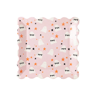 The Scatter Scallop Ghost Plate from My Mind’s Eye, a square pink paper plate with wavy edges adorned with a pattern of white ghosts, orange and black stars, and black triangular flags, is an ideal choice for a Halloween party.