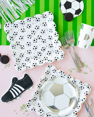 A table set with soccer-themed party supplies, including My Mind’s Eye Scattered Ball plates, cups, utensils, a napkin shaped like a soccer shoe, and cookies decorated to look like soccer balls. The background is green and white.