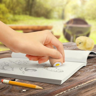 A hand uses a Fred and Friends Scribbled Eggs Erasers, which are BPA-free, to erase pencil drawings from an open sketchbook on a wooden table. Nearby, there is a yellow chick and a pencil. The erasers come packaged in a miniature egg carton. The background features a blurry outdoor setting.