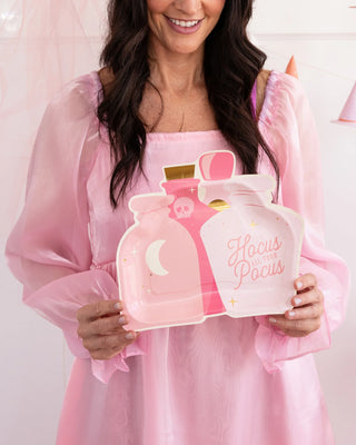 A woman in a pink dress holds the My Mind’s Eye Shaped Potion Plate with images of potion bottles and the text "Hocus Pocus," perfect for adding charm to your spooky festivities.