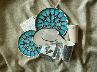 An assortment of party tableware with elegant turquoise patterns, including plates, cups, and Party West's Bride's Last Ride disposable cocktail napkins, artfully arranged on a textured beige fabric with silver fringe.