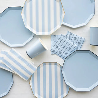 A minimalist table setting featuring geometrically shaped Sky Blue Signature Cabana Stripe Plates from Bonjour Fête, solid blue cups made from eco-conscious materials, and coordinating napkins on a light wooden surface.