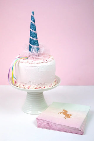 A white cake with a blue unicorn horn topper and rainbow ribbons, set on a white cake stand, next to Talking Tables Small Unicorn Napkins and other unicorn party supplies on a white surface with a pink background. Perfect for any kids party!