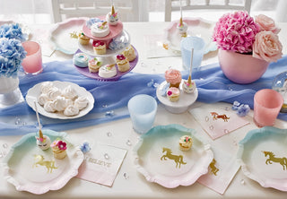 Decorated party table with unicorn-themed plates, cupcakes, meringues, pink and blue flowers, and glasses. Talking Tables Small Unicorn Napkins are placed around the setting with a blue table runner underneath—perfect for a magical kids party.