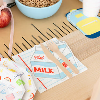 Table setting featuring Smarty Pants Milk Carton Napkins from Daydream Society, wooden fork and knife, apple, cereal bowl, and colorful cup coasters—ideal for a back-to-school breakfast or assembling party essentials.