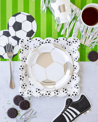 Soccer-themed table setup with My Mind’s Eye Soccer Ball Napkins, soccer ball plates, coasters, and more featuring soccer ball designs; a fork, a cup of tea, chocolate cookies, and a shoe-shaped decoration on a green and white background. Perfect for soccer-themed events or party decor.