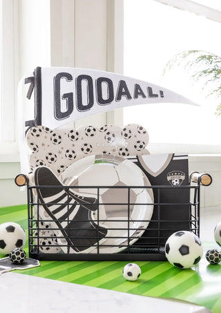 A wire basket filled with soccer-themed items, including My Mind’s Eye Soccer Ball Plates, napkins, a flag that reads "GOOAAL!", and mini soccer balls—perfect for soccer fans—placed on a green table near a window.