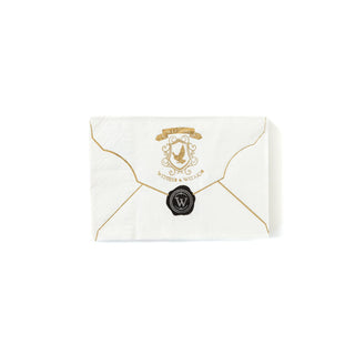 The Spellbound Envelope Napkin by My Mind’s Eye is a white napkin shaped like an envelope, adorned with golden edges. It features a crest with an 'M' and the words "Witches & Wizards" in shimmering gold, and is sealed with a black wax stamp.