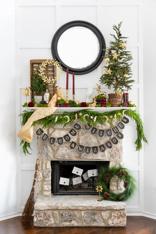 A rustic stone fireplace adorned with festive greenery, a round mirror, decorative candles, miniature Christmas trees, and a banner reading "Welcome Witches & Wizards." Spellbound Envelope Napkins from My Mind’s Eye and a wreath accompany the scene.