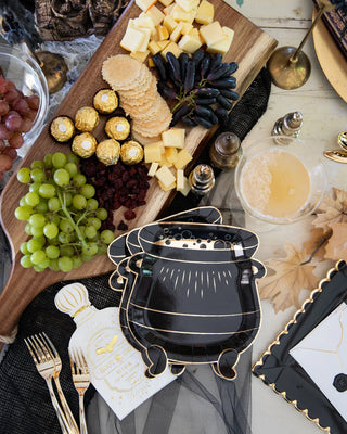 A festive table setting featuring a wooden board with grapes, cheese cubes, crackers, chocolates, and dried fruit, alongside a black cauldron-shaped plate, drinks, gold utensils from My Mind’s Eye, and Spellbound Potion Napkins.