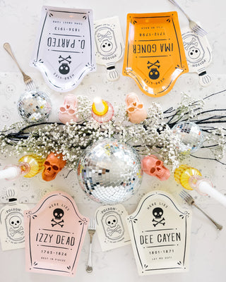 A Halloween-themed table setup with skull-shaped decorations, baby breath flowers, and gravestone-shaped plates labeled "D. Parted," "Ima Goner," "Izzy Dead," and "Dee Cayen" around a disco ball, complemented by My Mind’s Eye's Spellbound Potion Napkins for a touch of macabre magic.