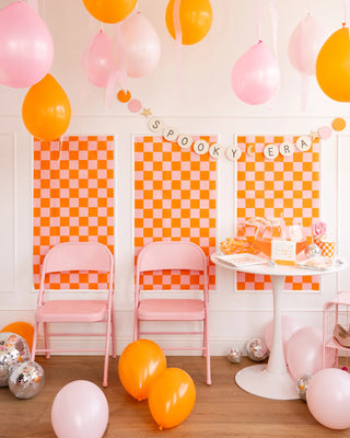 A decorated party room with pink and orange balloons, a My Mind’s Eye Spooky Era banner, orange and white checkered wall panels, two pink foldable chairs, gold glitter accents, and a white table with party supplies.