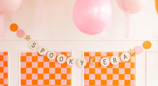 The "Spooky Era" banner by My Mind’s Eye adds a festive touch to your Halloween party backdrop, featuring an orange and pink checkerboard design complemented by hanging pink balloons and gold glitter accents.