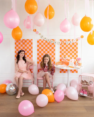 Two children in pink outfits sit amidst pink and orange balloons with a "Spooky Era" banner from My Mind’s Eye, set against checkered orange backgrounds, creating a festive atmosphere. Gold glitter accents add a touch of shimmer to the Halloween party decoration.