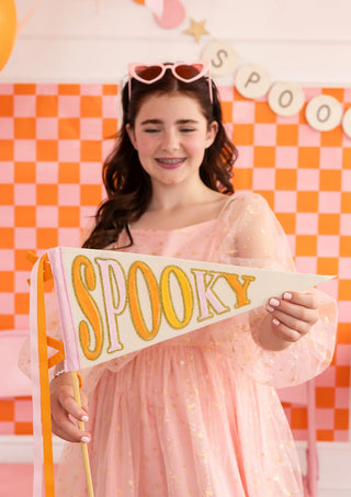 A girl in a light pink dress holding My Mind's Eye's Spooky Felt Pennant. There is a pink and orange checkered background, adorned with a Halloween-themed banner spelling "SPOOKY" that adds to the festive ambiance.