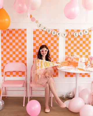 A person in a pink dress and heart-shaped glasses sits on a pink chair at a party with pink and orange decorations, holding the "Spooky Felt Pennant" by My Mind’s Eye. The backdrop features a Halloween-themed banner reading "Spooky Era.