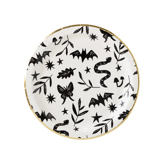 The Spooky Icons Plate by My Mind’s Eye is perfect for a Halloween party, featuring black illustrations of bats, snakes, mushrooms, plants, and stars on a white background with a gold rim.