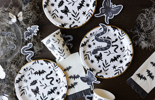 A Halloween-themed table setup features the Spooky Icons Plate by My Mind’s Eye, showcasing snake, bat, and spider designs on paper plates, cups, and napkins. Halloween decorations such as bats and spiders are scattered around for a truly spooky celebration.