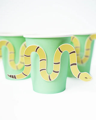 Three light green paper cups, each adorned with a yellow and brown snake illustration that wraps around the cup, perfect for your cowboy party. These Snake Catcher Cowboy Party Cups by Pop! Party Supplies are a standout addition to any western party.