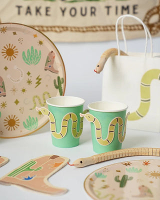 Table setup featuring snake-themed paper plates, Snake Catcher Cowboy Party Cups from Pop! Party Supplies, and a toy snake. Cactus and cowboy boot illustrations also adorn some items. A cloth bag from the snake catcher collection and a backdrop with the text "TAKE YOUR TIME" are visible, perfect for a western party.