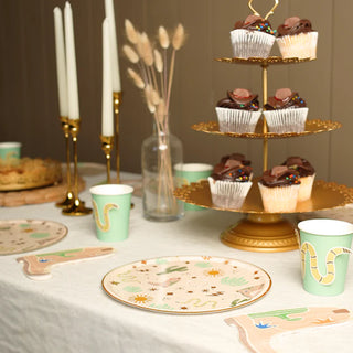 A table set for a Western Party with cupcakes on a tiered stand, themed plates and cups featuring designs with snakes, Snake Catcher Napkins by Pop! Party Supplies, and two lit candles in golden holders.