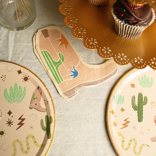 A table decorated with a cowboy boot-shaped napkin, Pop! Party Supplies Snake Catcher Western Theme Paper Plates featuring cactus designs, cupcakes, and a scalloped edge tray adorned with gold foil details.