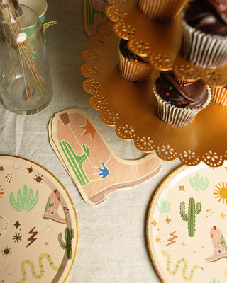 Table setup with Snake Catcher Western Theme Paper Plates by Pop! Party Supplies featuring gold foil details, a napkin shaped like a cowboy boot, a tiered cupcake stand with chocolate cupcakes, and a glass containing skewers. Perfect for your next cowboy party!