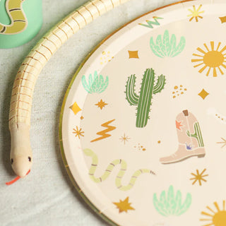 A round plate with desert-themed illustrations, including cacti, a cowboy boot, a snake, stars, and a sunburst. The Pop! Party Supplies Snake Catcher Wooden Snake is placed next to the plate on a light surface, perfect for customizable decorations or party favors.