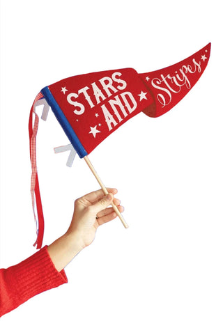 A person in a red sleeve waves a patriotic 'My Mind's Eye Stars and Stripes' felt pennant banner against a backdrop of vertically hanging red, white, and blue stripes, invoking American national pride.