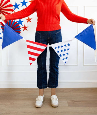 A person wearing a red sweater, blue jeans, and white shoes holds a My Mind's Eye Stars and Stripes Outdoor Banner with stars and stripes, standing in front of a wall decorated with red, white, and blue.