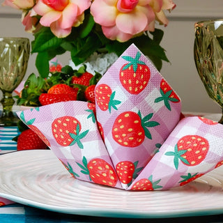 A folded Strawberry Gingham Paper Napkin from Talking Tables with a strawberry design is placed on a white plate. In the background, perfect for picnics in the park, strawberries, flowers, and green glassware are visible. Plus, these napkins are 100% recyclable!