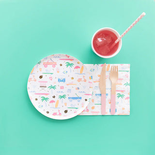 A Surf's Up Large Plate from Daydream Society and a napkin with colorful prints sit alongside a wooden fork and knife featuring pastel handles. Completing the scene is a cup containing a pink drink with a straw, all arranged on a turquoise background that perfectly captures the essence of beachside surf culture.