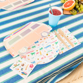 Table setting with a striped tablecloth featuring Daydream Society's Surf's Up Large Plates, accompanied by matching napkins. A blue cup with a pink drink and a fruit platter embody the vibrant energy of beachside surf culture nearby.