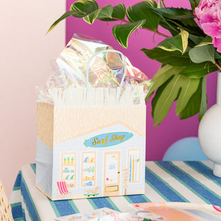 The "Surf's Up Party Bag" by Daydream Society rests on a striped tablecloth, encircled by vibrant green and pink plants, embodying the spirit of beachside surf culture.